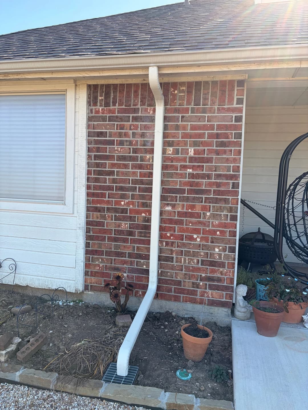 Downspout attached to brick wall, directing rainwater into drain near potted plants.