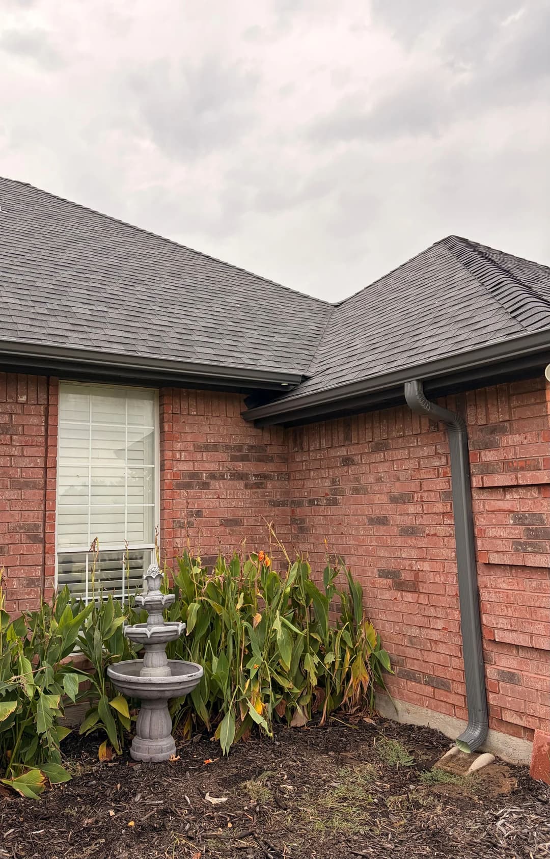 Brick house corner with a decorative fountain and lush green plants under a cloudy sky.