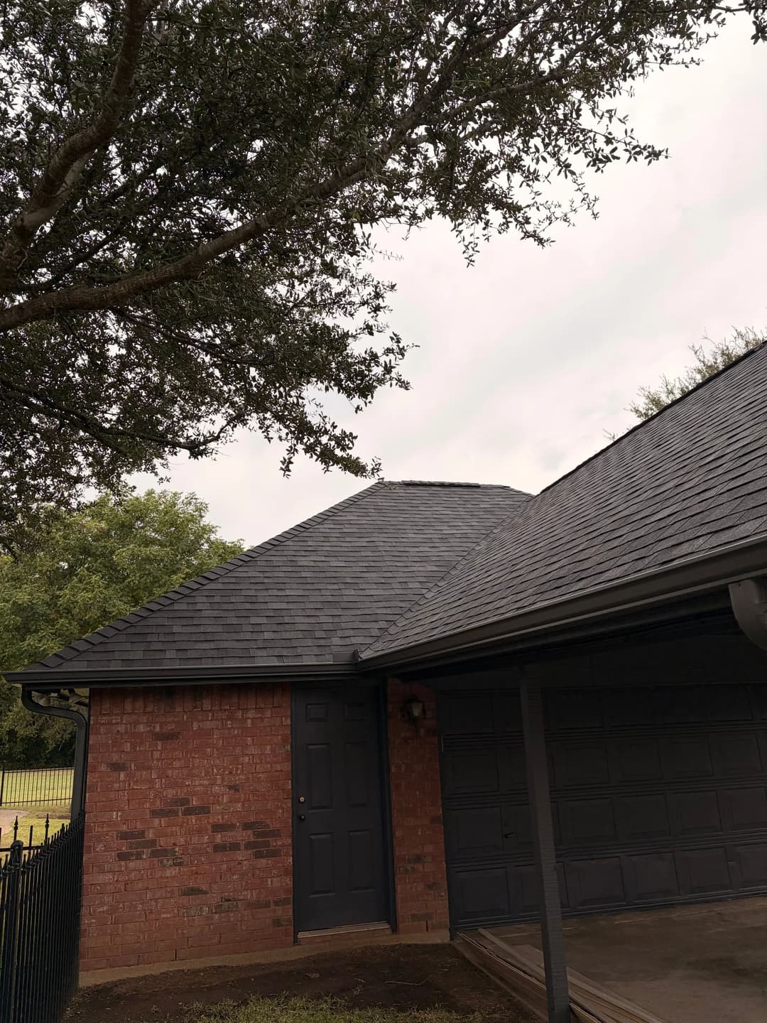 Brick house with a dark shingle roof and a large tree, set against an overcast sky.