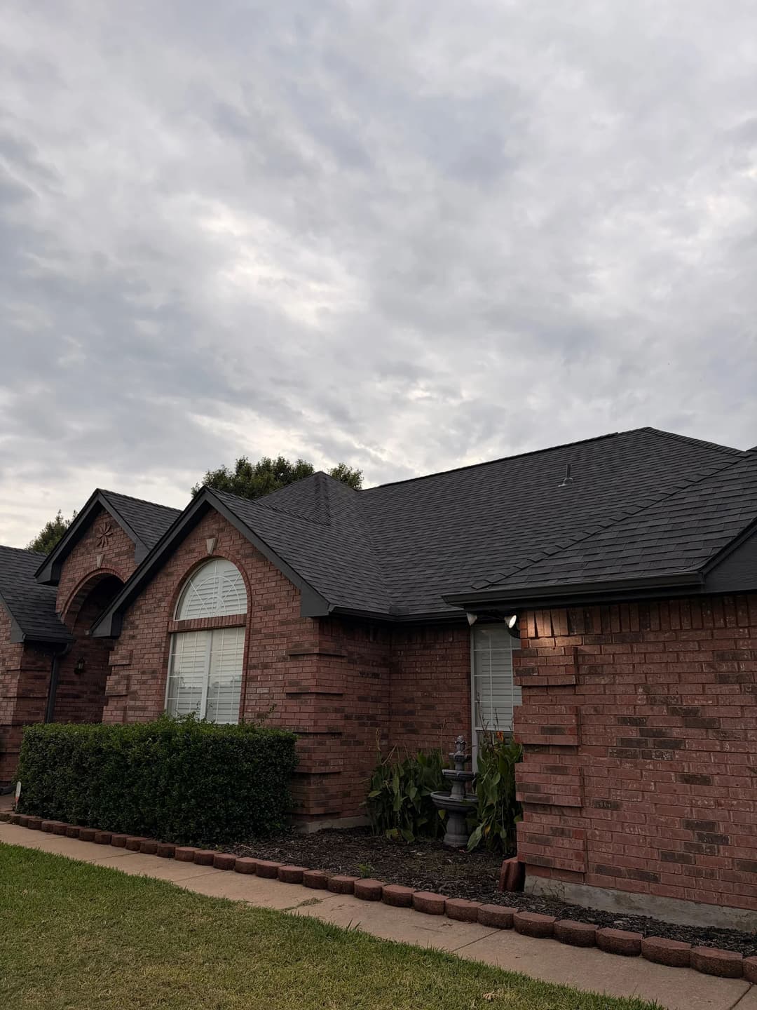 Brick house with a dark roof, green bushes, and cloudy sky in the background.